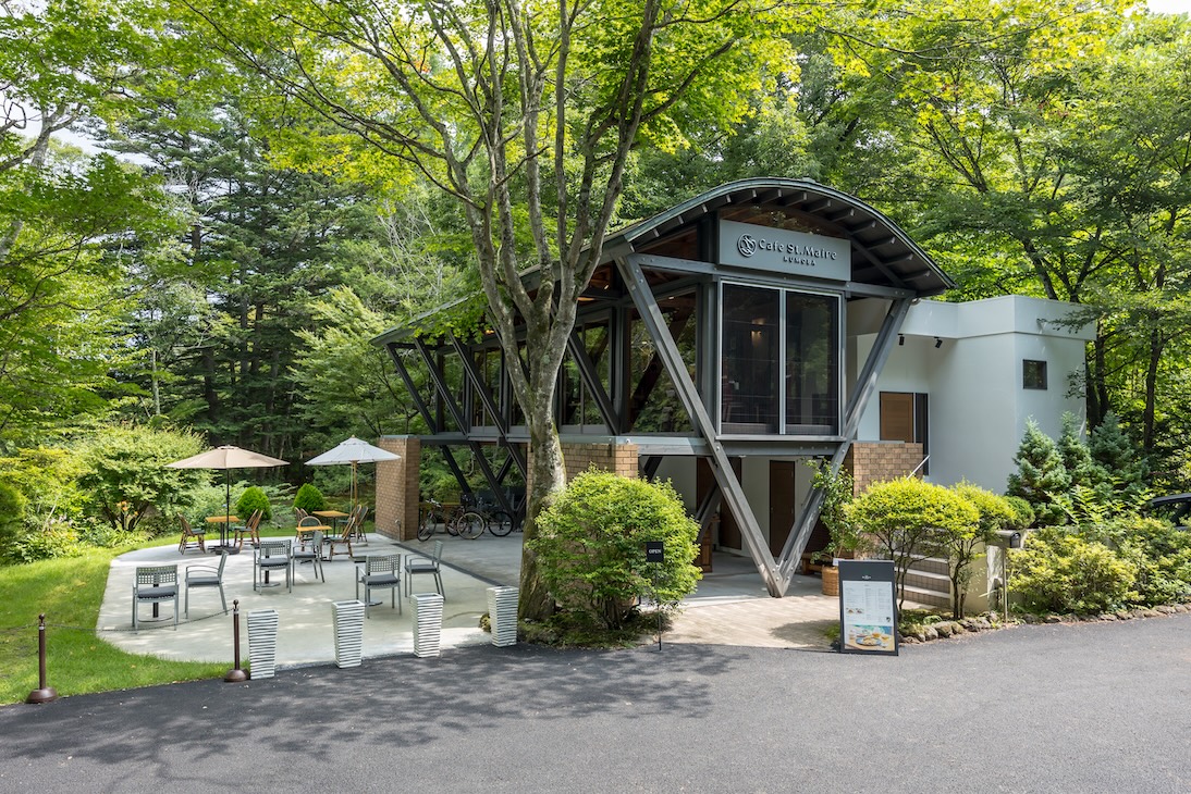 「Cafe St.Maire KUMOBA（カフェ サンメール雲場）」の外観。緑豊かな森の中に佇む建物で、大きなガラス窓と特徴的な三角形の柱が印象的です。建物の前にはテラス席があり、パラソルの下にテーブルと椅子が設置されています。自然と調和したデザインが魅力です。