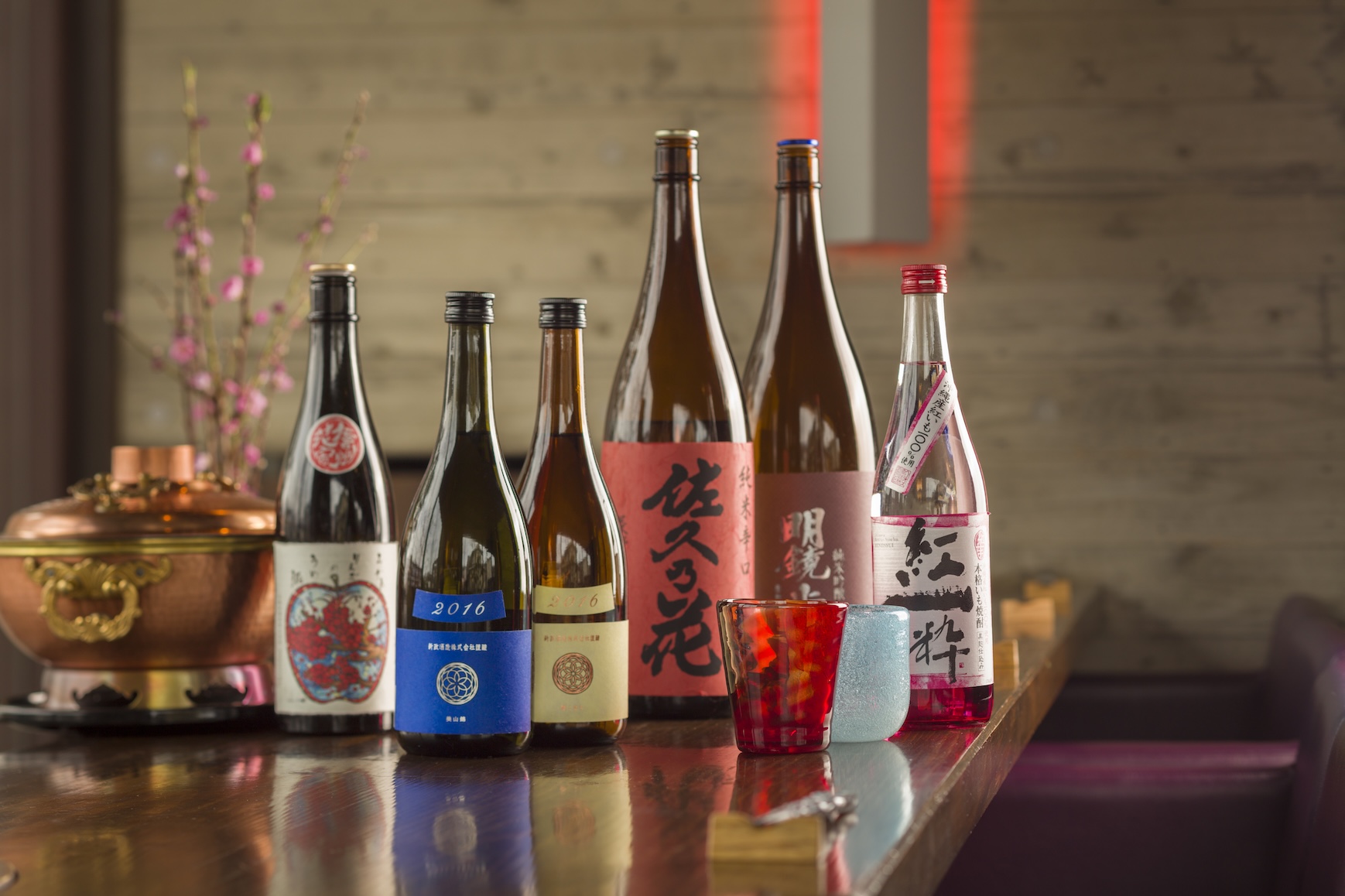 テーブルに並べられた日本酒の瓶とカラフルなグラス。6本の日本酒の瓶は異なるラベルデザインで、それぞれ個性的な銘柄を表しています。手前には赤と青のガラス製のグラスが置かれ、奥には銅製の鍋と背景に梅の枝が飾られた落ち着いた和風の雰囲気が漂います。高級感と伝統美を感じさせる場面です。